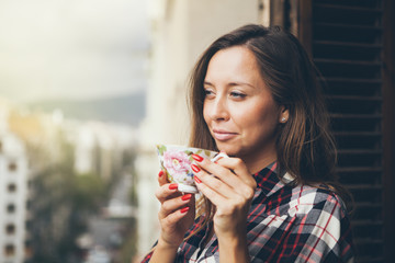 Woman is smiling and drinking coffee on a balcony with city view. Close up of a woman holding in hands and cup of coffee. Early morning routine. Sun light flair effect