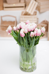 Beautiful light rosy tulips in glass vase on white table, close up. Variety of defocused wooden boxes on background. Floristics, arranging flowers for presents and decoration concept