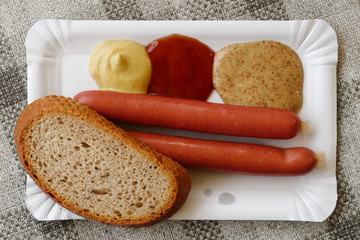 Pair of frankfurter sausages with a slice of bread on paper plate from above. Grey tablecloth.