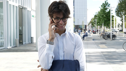 Fototapeta premium Handsome young businessman in city talking on cell phone, also looking at camera, serious
