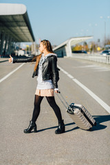 Fuul body vertical photo of young hipster woman with suitcase hitch-hiking on the road in the city background. Concept of travel, holidays, journey, trip.