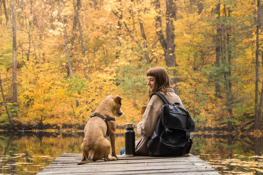 Girl Hiker Rests At Riverbank With Her Pet Dog And Drinks Coffee From Thermos. Young Female Person And Her Puppy Having A Rest By The Lake On Beautiful October Day Of Indian Summer