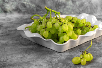 Branch of a green grapes on a white plate on a gray marble background with copy space and selective focus