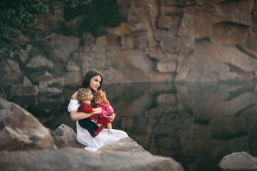 A beautiful model-looking girl is sitting by the river and carefully holding two dolls, similar to children, in her arms. A young woman imagines that she is a mother.