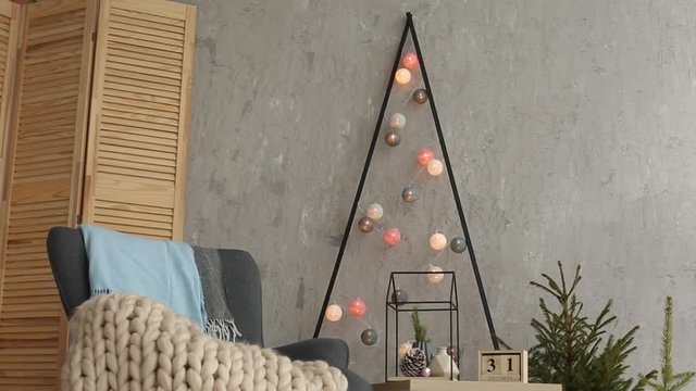 Stylish christmas scandinavian interior with an elegant armchair and warm merinos plaid. Comfort home with nordic new year decor. Minimalistic christmas tree with garlands and lights in light room.