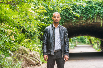 Portrait of young American hispanic man. Young man with short hair wearing gray shirt, black leather jacket, black pants, standing in front of street bridge with green plants at Central Park, New York