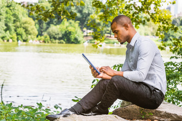 Fototapeta premium Young American hispanic man traveling, working in New York. Young guy with short hair wearing gray shirt, black pants, leather shoes, holding tablet computer, sitting by lake at Central Park, reading