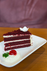 Slice of homemade velvet red cake  decorated with cream on white dise and wood table