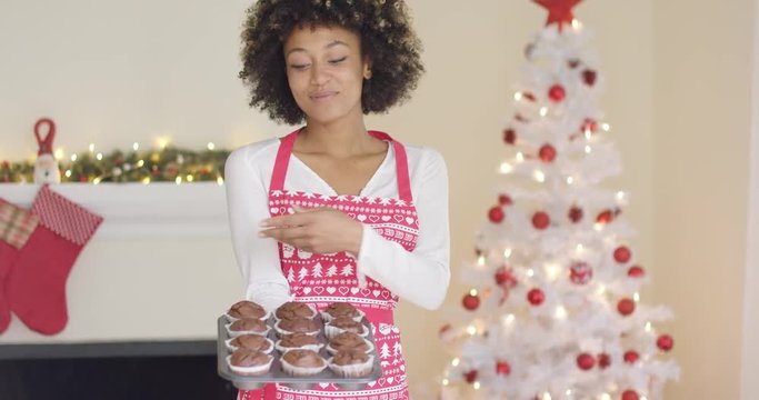 Proud young cook displaying her freshly baked tray of Christmas muffins as she stands in her apron in front of the tree