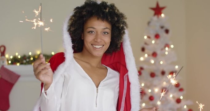 Smiling young woman in a Santa Claus cloak celebrating Christmas at home in front of the tree holding a burning sparkler