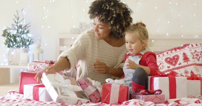 Mother showing cute little child a large red gift box for Christmas while seated on bed in beautifully decorated bedroom