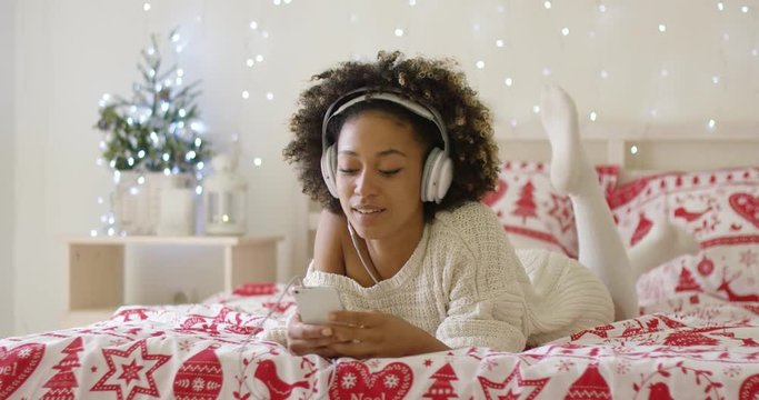 Attractive young woman relaxing at Christmas lying on her bed listening to music on her mobile with sparkling festive lights in the background