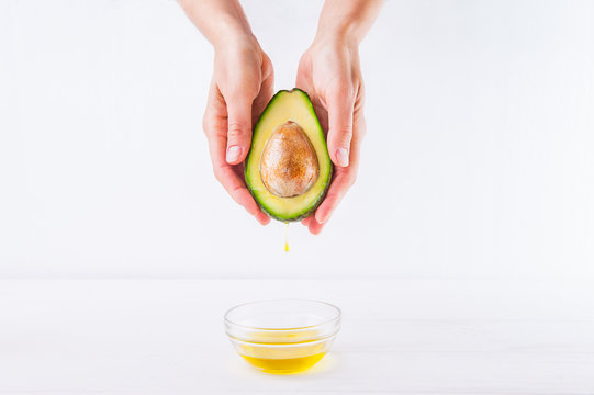 Fresh Avocado With Oil Drop In Female Hands And Bowl Of Oil On The White Wooden Background Isolated. Healthy, Natural Beauty Concept. Front View. Space For Text.