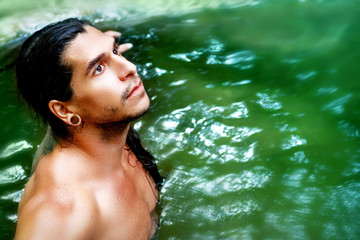 A handsome guy with long hair and piercings on waterfalls in a rain forest against a background of green water.