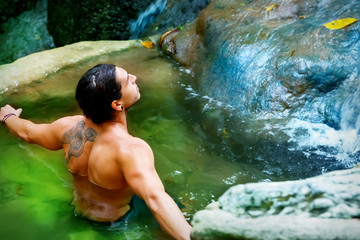  Нandsome guy with long hair and piercings on waterfalls in a rain forest against a background of clear blue and green water.