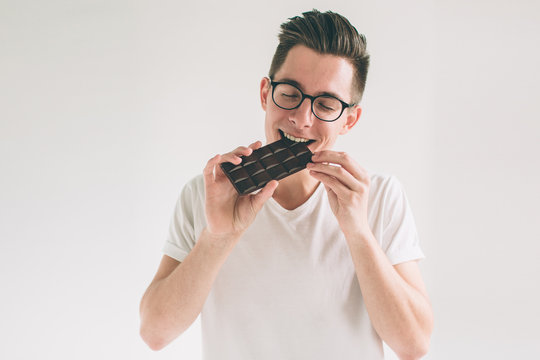 young man eating a chocolate bar. Nerd is wearing glasses.