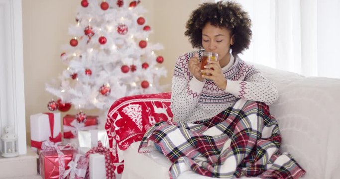 Young woman relaxing at home over Christmas sitting under a warm rug on the sofa with a cup of tea in front of the tree
