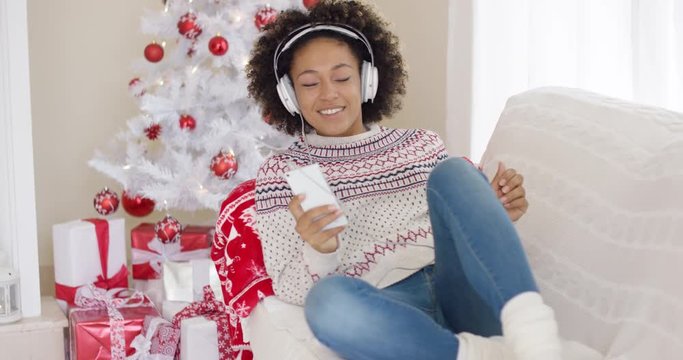 Young girl relaxing on a sofa in front of the decorated tree listening to Christmas music on her mobile phone on stereo headphones