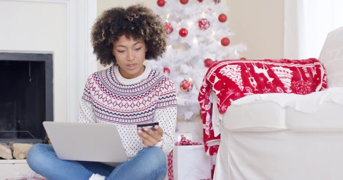 Young woman purchasing Christmas gifts online as she sits cross legged on the floor in front of the decorated tree at home entering her credit card details on a laptop.