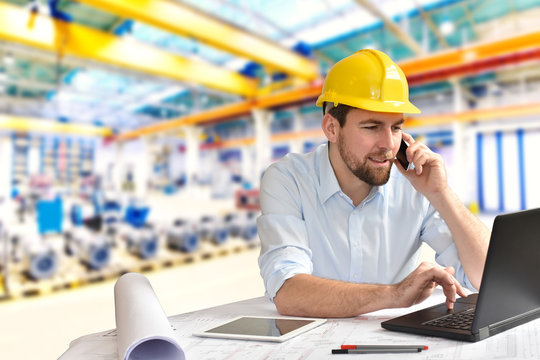 Ingenieur im Maschinenbau telefoniert am Arbeitsplatz/ Schreibtisch in einer Fabrikhalle und arbeitet am Rechner