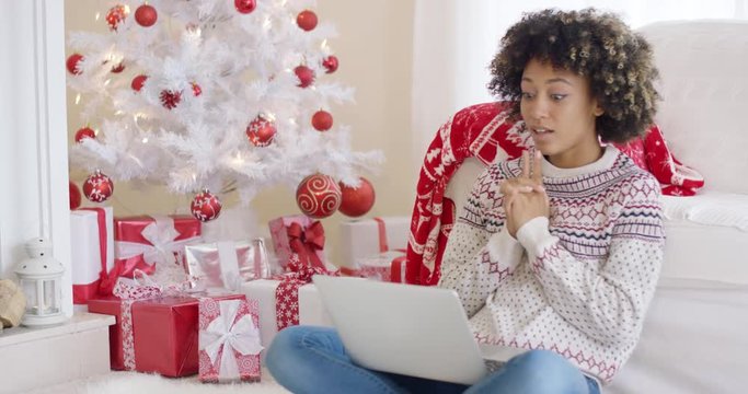 Young woman typing on a laptop at Christmas sitting on the floor in front of the tree and a pile of colorful red and white gifts.