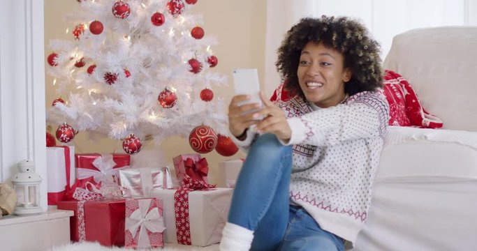 Young woman photographing her Christmas tree on her mobile phone to share with friends on social media sitting on the floor pointing to it