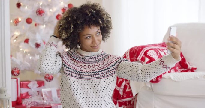 Happy young woman posing for a Christmas selfie on her mobile phone in front of a red and wite themed tree and gifts in a festive living room.