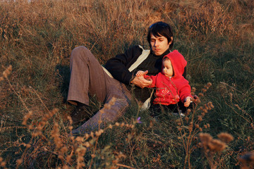 Naklejka na ściany i meble Happy family father and child daughter on nature on sunset. A child of one two years and a parent are sitting in a high dry grass on a meadow. Autumn. daughter and father are looking at the grass