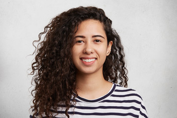 Headshot of curly mixed race beautiful female with smile wears striped sweater, isolated over white background. Caucasian young woman has crisp dark hair and appealing appearance. Beauty concept