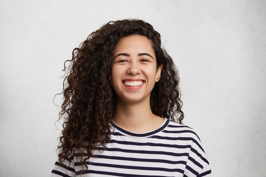 Portriat of pretty curly female wears striped black and whitte t shirt, smiles happily as being glad to meet best friend, who tels her funny jokes or stories. Happiness and facial expressions concept