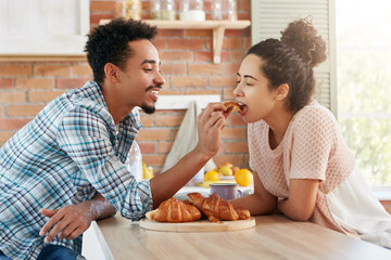 Affectionate bearded mixed race man feeds his girlfriend with tasty croissant which he baked by himself. Adorable curly woman closes eyes with pleasure as tastes delicious cake, sits in kitchen