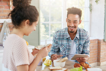 Indoor shot of happy bearded man with specific appearance, uses tablet computer for searching social networks, eats delicious dishes prepared by his hard working wife. Couple have talk at kitchen
