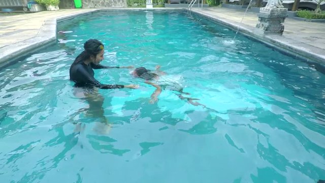 Young woman teaching her daughter to swim while wearing swimsuit and swimming goggles at pool with blue water