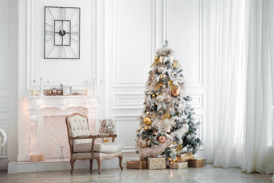 Classic white christmas interior with new year tree decorated. Fireplace with grey chair, clocks on the wall and presents under the tree