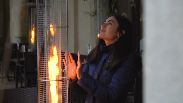 Girl brunette warms her hands near the flame of street gas heaters