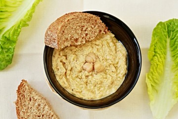 Top view of homemade hoummus with chickpeas on top in a black bowl with pieces of wholemeal bread and bright green leaves of lettuce