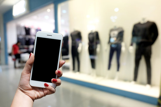 woman holds a smartphone standing in front of a mall. There is a blurred image of the clothing store as the background.