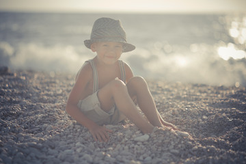 Cute old style boy kid on beach walking posing wearing fancy shorts with gallows and gentleman hat enjoy summer time alone on awesome ocean with anchor.Adorable scene of childhood life - obrazy, fototapety, plakaty