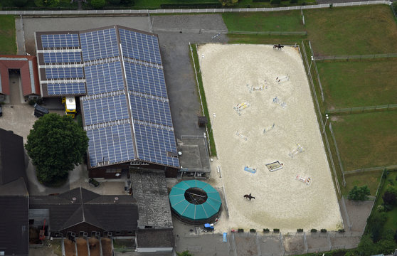 Luftaufnahme des mit Solarzellen belegten Dachs einer Reithalle mit nebenliegendem Reitplatz mit zwei Reitern