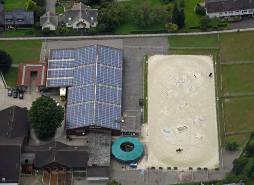 Luftaufnahme des mit Solarzellen belegten Dachs einer Reithalle mit nebenliegendem Reitplatz mit zwei Reitern