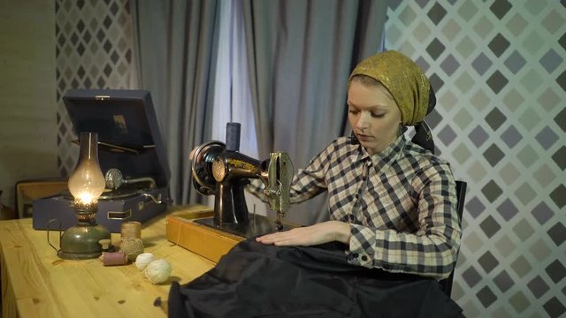 Retro seamstress girl sews cloth with old manual hand sewing machine. Woman working at home or workshop at night with kerosene lamp, listens music vinyl plate, gramophone or phonograph