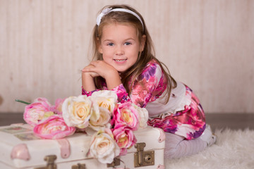 Cute baby girl with brunette hairs pink cheeks and green eyes posing sit for camera in studio wearing airy colourful dress and fancy casuals pumps close to travelling trunk bag with pictures and roses