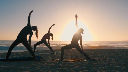 Group of 3 women practicing yoga on the beach against the sunset. Three black silhouettes of their bodies making yoga postures. Sun and the ocean creates ethereal & pure scene