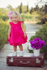 Amazing deep blue eyes baby girl child stylish dressed in colourful pink dress with shining blond hairs and white sandals posing sit for camera summer central park forest meadow with flowers and trunk