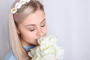 Obraz na płótnie Canvas Beautiful young blonde woman with clean skin and flower wreath in her hair smelling bouquet white rose, eyes closed