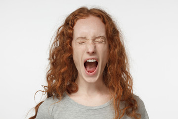 Closeup portrait of screaming with closed eyes crazy redhead curly girl in grey t-shirt isolated on gray background