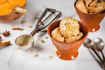 Ideas for autumn desserts, recipes from pumpkins. Pumpkin pie ice cream gelato in ceramic bowls, with maple syrup, pumpkin seeds, cinnamon and anise stars, on a white marble table. Copy space