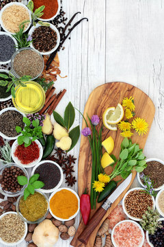 Herbs, spices and edible fresh flowers with olive oil in bowls and loose on an olive wood board with knife forming a colourful rustic abstract background. Top view.