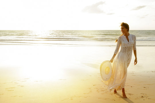 Woman wearing beautiful white dress is walking on the beach during sunset