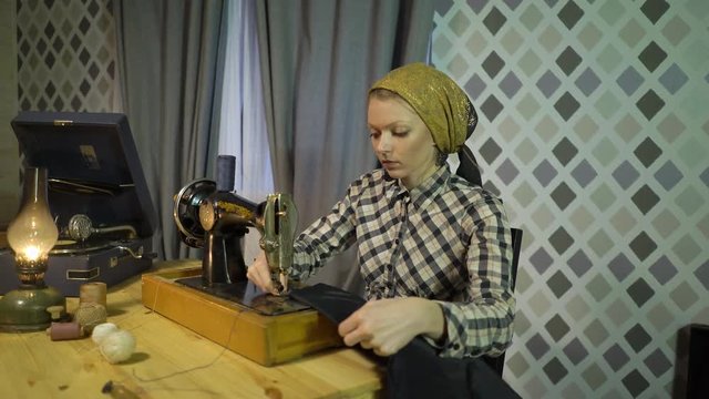 Beautiful retro seamstress girl sews cloth with old manual hand sewing machine. Woman working at home or workshop at night with kerosene lamp, listens music vinyl plate, gramophone or phonograph